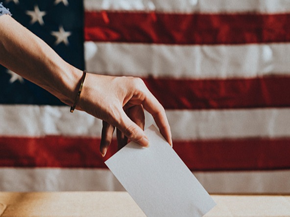 Hand places vote in ballot box with a US flag in the background